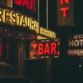 Night street scene with a glowing yellow 'RESTAURANT' sign and a red-lit 'BAR' sign beneath, in a dark urban storefront at night.