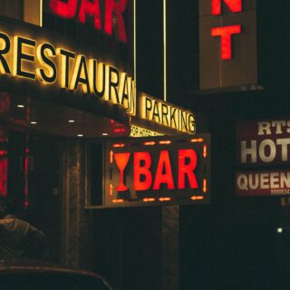 Night street scene with a glowing yellow 'RESTAURANT' sign and a red-lit 'BAR' sign beneath, in a dark urban storefront at night.