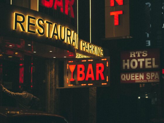 Night street scene with a glowing yellow 'RESTAURANT' sign and a red-lit 'BAR' sign beneath, in a dark urban storefront at night.