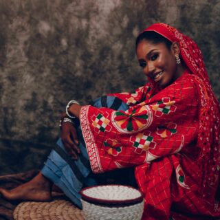Woman in a bright red patterned garment sits cross-legged beside a woven basket on a dark backdrop.