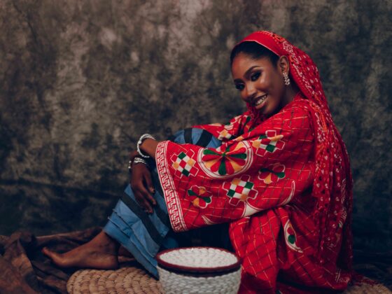 Woman in a bright red patterned garment sits cross-legged beside a woven basket on a dark backdrop.