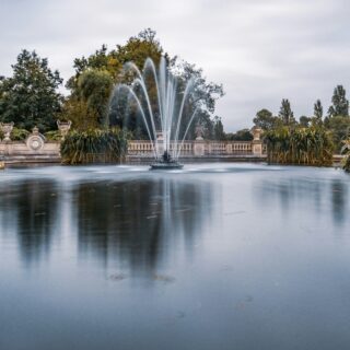 Ornate fountain in a pond with a stone balustrade and surrounding greenery in a formal garden park.
