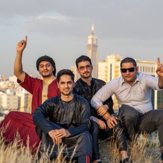 Group of four men posing on a grassy hill with a city skyline behind them; two point upward with one raised finger gesture.