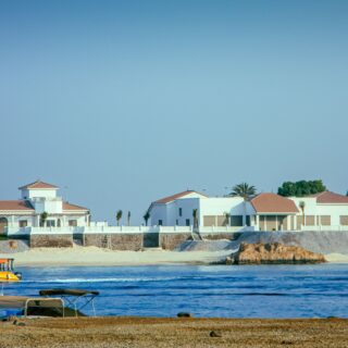 Coastal row of white houses with red-tiled roofs along a marina, calm blue water in the foreground and boats docked nearby.