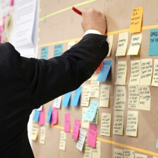 Person in a dark suit places sticky notes on a planning wall covered with colorful notes and markers in use.