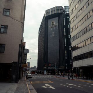 City street lined with tall modern buildings; pedestrians cross at a crosswalk and cars wait at a light in a busy urban district.