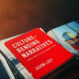 Close-up of a bright red book cover reading 'Culture-Bending Narratives' by Jason Lacy, with a photo of buildings on the right edge.