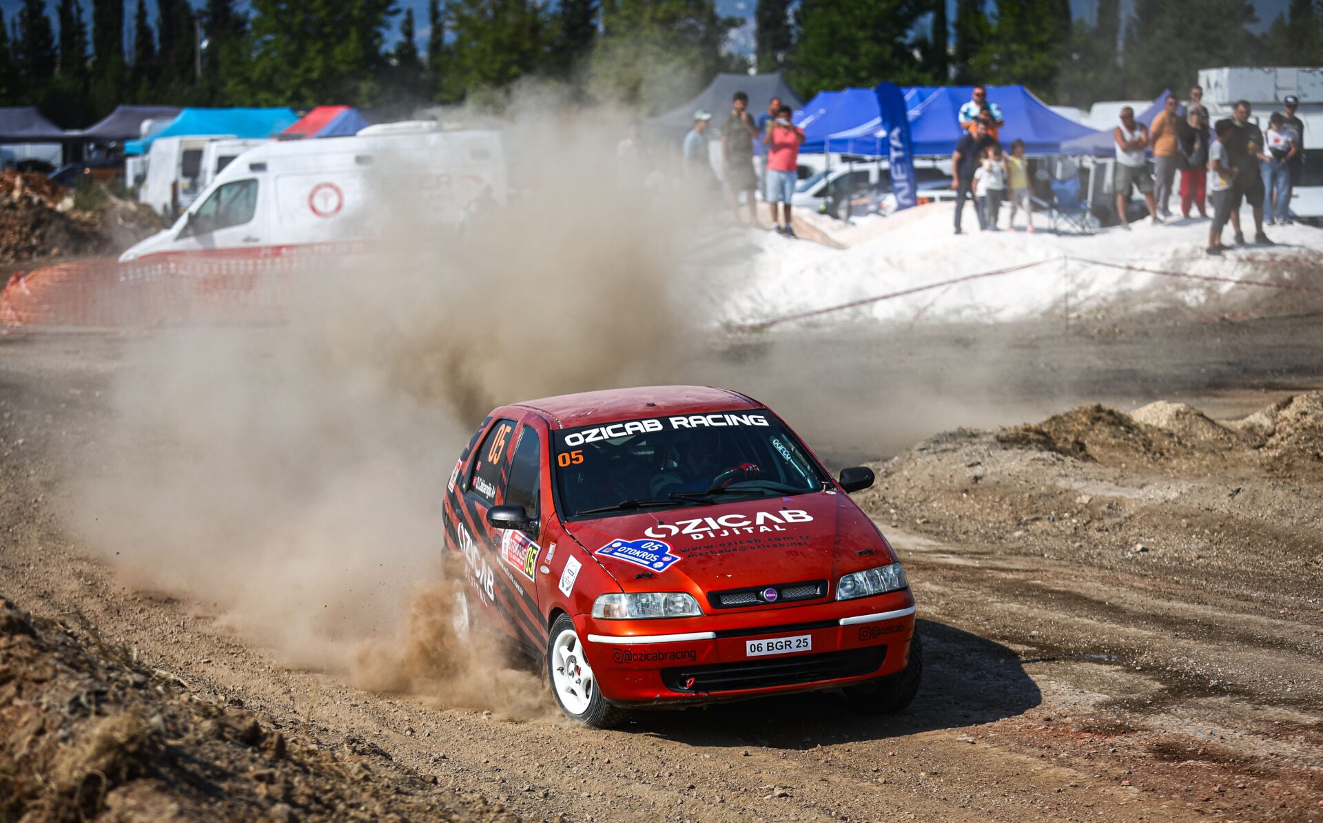 Red rally car number 05 speeding on a dirt track, kicking up dust as spectators and tents watch nearby.
