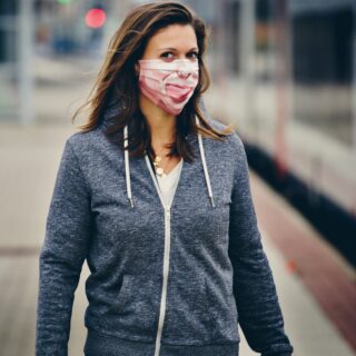 Woman wearing a pink patterned face mask and gray zip hoodie at a train platform, looking toward the camera with a blurred background.