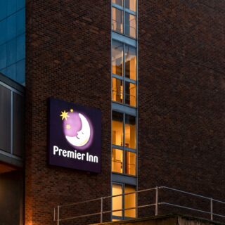 Brick building at dusk with a vertical row of lit windows and a glowing Premier Inn sign outside.
