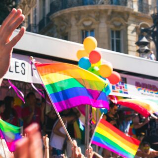 Crowd at a Pride parade waving rainbow flags and balloons, hands raised in celebration with city buildings in the background.