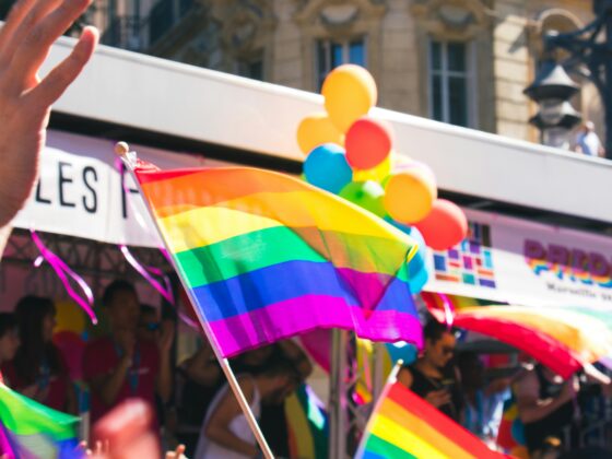 Crowd at a Pride parade waving rainbow flags and balloons, hands raised in celebration with city buildings in the background.