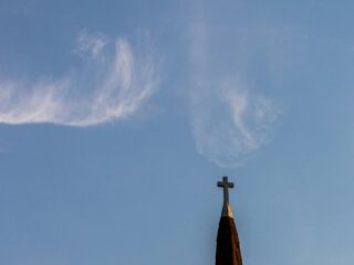 Church steeple with a cross at the top against a clear blue sky with wispy clouds (lower right).