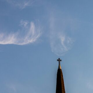 Church steeple with a cross at the top against a clear blue sky with wispy clouds (lower right).