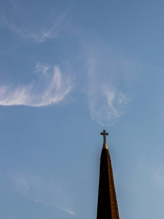 Church steeple with a cross at the top against a clear blue sky with wispy clouds (lower right).