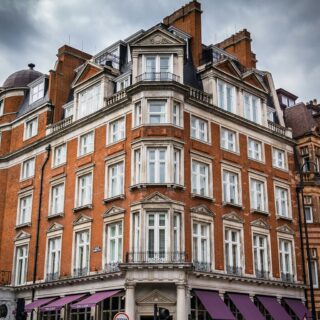 Red brick, ornamented city buildings under a cloudy sky; pedestrians stand along the sidewalk in front of purple-awning shops laterally.