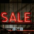 Red neon letters spell 'SALE' against a dark wooden backdrop in a storefront window.