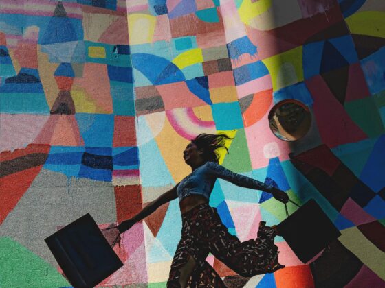 Person wearing patterned pants and boots walks along a vivid geometric mural, with a round mirror on the wall to the right.
