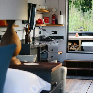 Interior of a modern camper van kitchen with stainless sink, dish rack, kettle, and red fire extinguisher on a shelf, open door to greenery outside