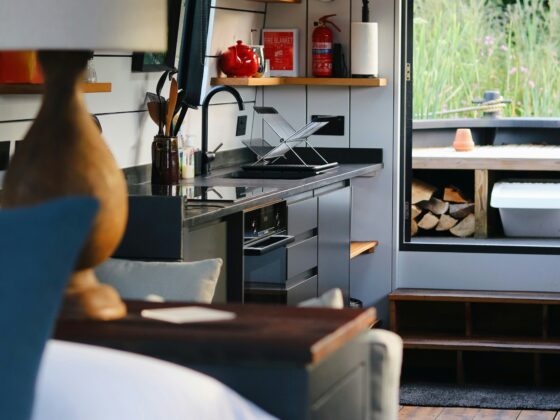 Interior of a modern camper van kitchen with stainless sink, dish rack, kettle, and red fire extinguisher on a shelf, open door to greenery outside