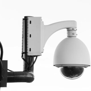 Black-and-white photo of a street lamp mounted on a pole with cables and a gear-like mounting bracket on the left side.