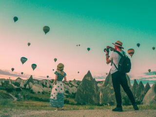 Two people on a rocky field photographing dozens of colorful hot air balloons rising into a pastel sky.