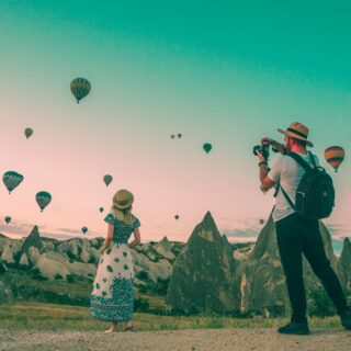 Two people on a rocky field photographing dozens of colorful hot air balloons rising into a pastel sky.