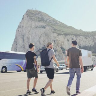 Three casually dressed men walking along a road beside a tour bus with a rocky mountain in the background.