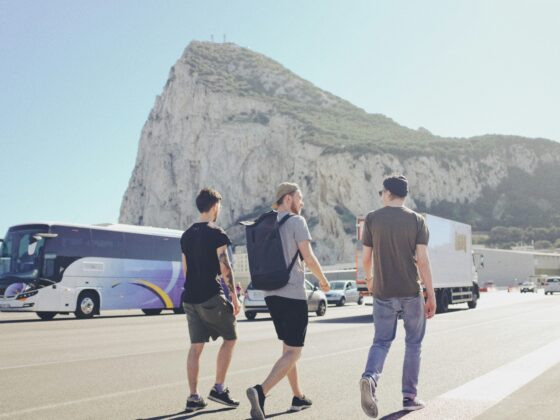 Three casually dressed men walking along a road beside a tour bus with a rocky mountain in the background.