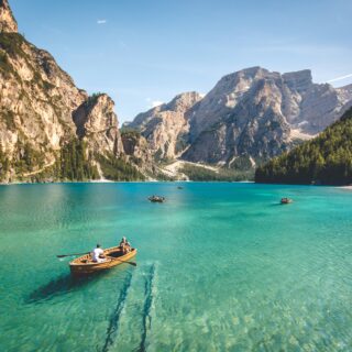 Turquoise alpine lake surrounded by rocky mountains and forest; several small wooden boats with people swimming and paddling near shore.