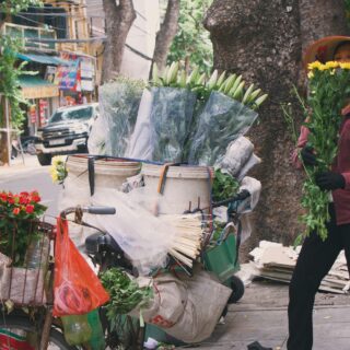 Street florist on a bicycle cart with buckets of flowers and yellow blooms on a busy urban street, vendor wearing a conical hat and red check shirt.