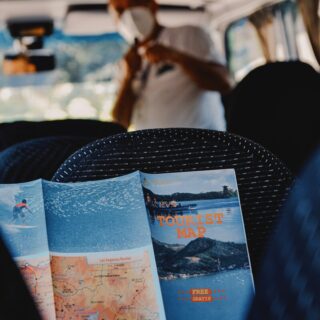 Tourist map booklet held by a passenger on a bus, with a cover reading 'TOURIST MAP' and a map of routes visible on the page in the foreground .
