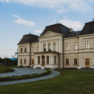 Beige historic building with ornate white trim, three steep dark roofs, and a curved cobblestone driveway against a clear blue sky.