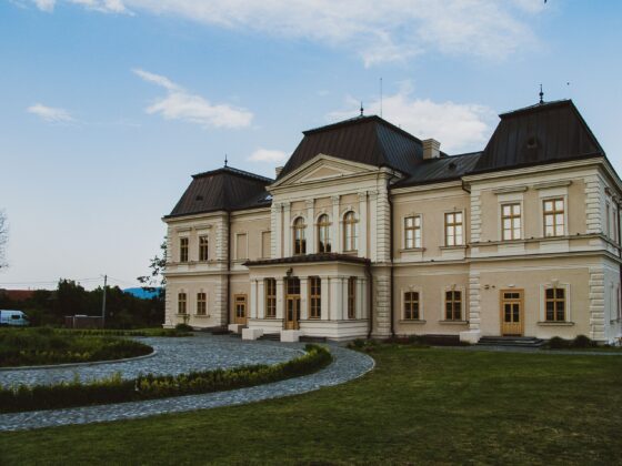 Beige historic building with ornate white trim, three steep dark roofs, and a curved cobblestone driveway against a clear blue sky.