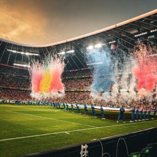 Soccer stadium celebration with colorful smoke bursts (red, yellow, blue) across the field and players lined up along the sideline.