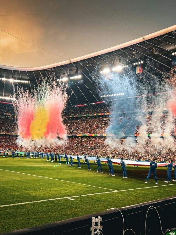 Soccer stadium celebration with colorful smoke bursts (red, yellow, blue) across the field and players lined up along the sideline.