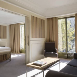 Sunlit hotel suite living area with a black sofa, wooden coffee table, beige curtains, and a balcony; doorway to a bedroom with a bed.