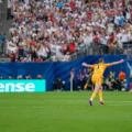 Soccer goalkeeper in an orange kit with pink gloves celebrating with arms outstretched on a green field, crowd cheering in the stands behind a Hisense signboard.