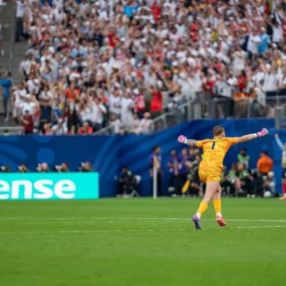 Soccer goalkeeper in an orange kit with pink gloves celebrating with arms outstretched on a green field, crowd cheering in the stands behind a Hisense signboard.