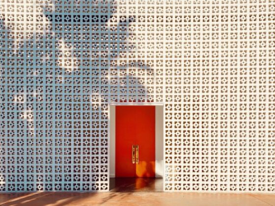Perforated white wall with square grid pattern and a red double door in the center; a person's shadow is cast across the wall above the door.