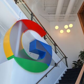 Oversized Google logo sculpture in primary colors mounted on a white wall beside a staircase in a modern office.