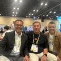 Three men at a trade show booth posing for a photo, wearing conference badges and lanyards.