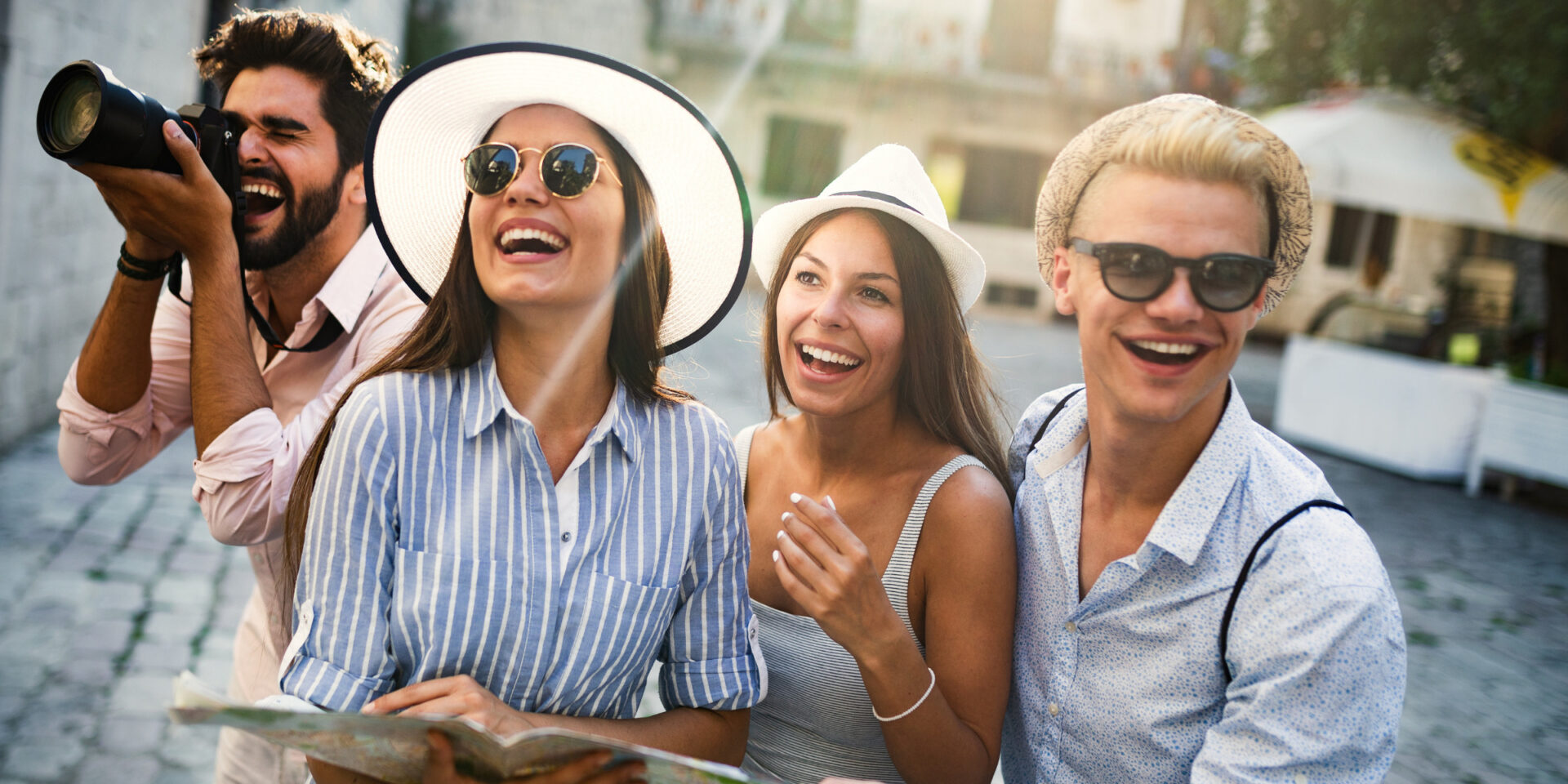 Four friends posing for a sunny street photo; one holds a camera while the others smile and laugh.