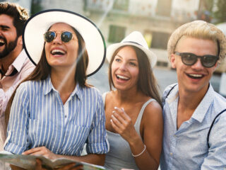 Four friends posing for a sunny street photo; one holds a camera while the others smile and laugh.