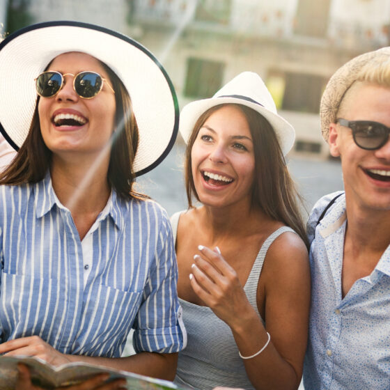 Four friends posing for a sunny street photo; one holds a camera while the others smile and laugh.