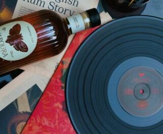 Blue vinyl record on a turntable with scattered photos, papers, and a whiskey bottle in a flat lay composition.