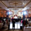 Elegant ballroom scene with a DJ booth on a raised stage, crystal chandeliers, and guests seated at round tables enjoying a formal reception.