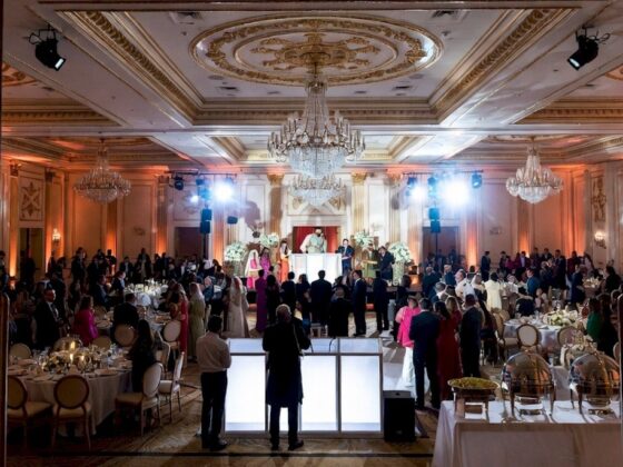 Elegant ballroom scene with a DJ booth on a raised stage, crystal chandeliers, and guests seated at round tables enjoying a formal reception.