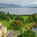 A lakeside estate with a castle-like hotel, manicured lawns, and a seaplane docked near the shore.