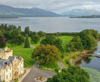 A lakeside estate with a castle-like hotel, manicured lawns, and a seaplane docked near the shore.